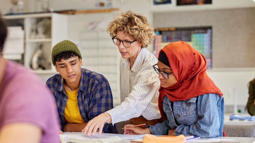 Lehrkraft unterstützt zwei Schülerinnen und Schüler am Tisch in einem hellen Klassenzimmer