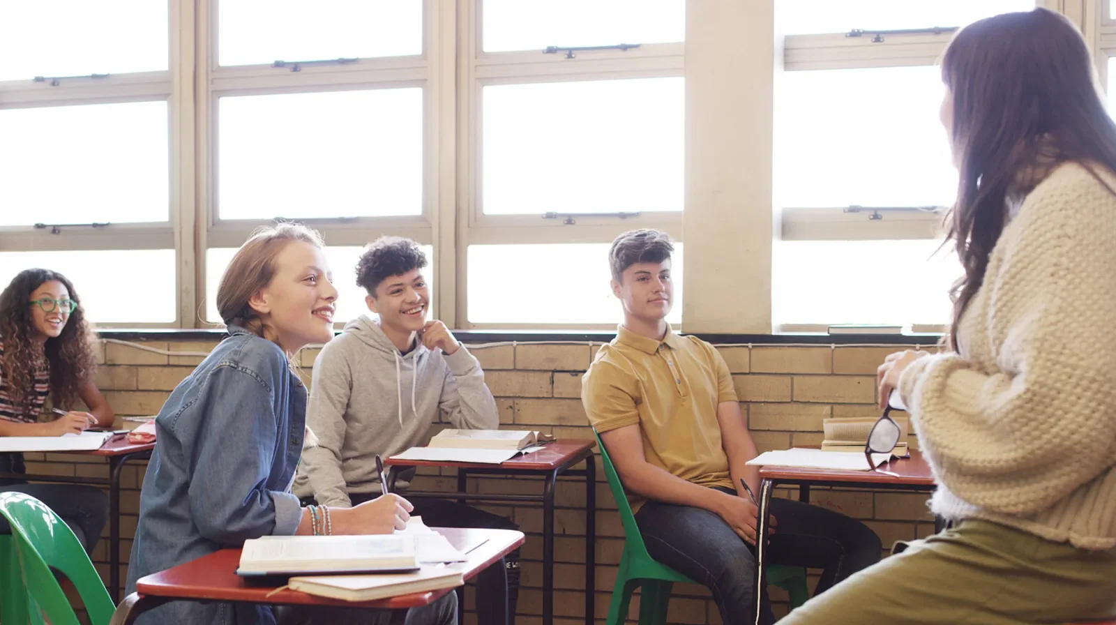 Lehrerin sitzt auf der Tischkante, hält ihre Brille in der Hand und blickt zu ihren Schülerinnen und Schülern, die ihr zuhören.